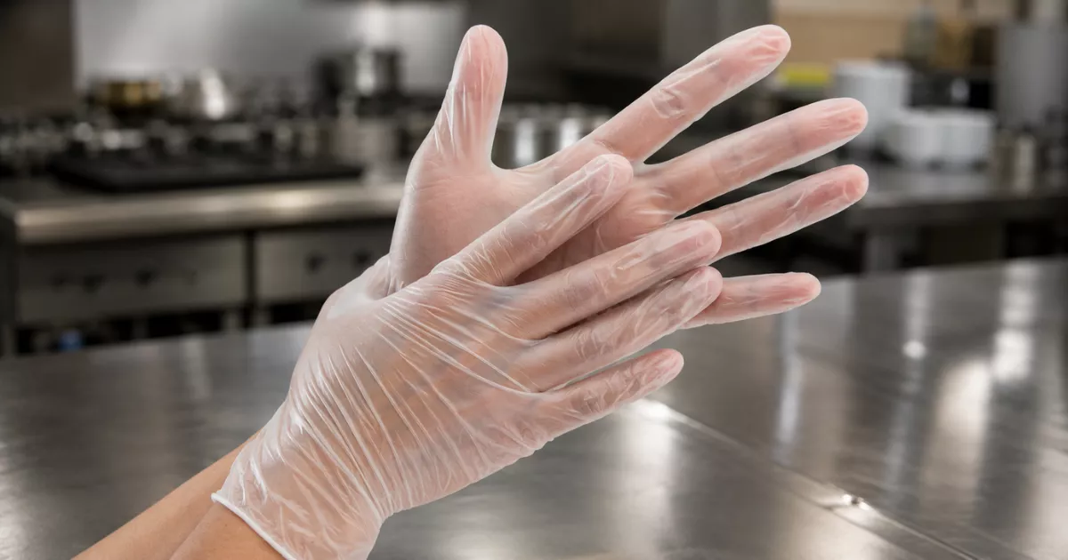 Duas mãos calçando luvas descartáveis de vinil transparente sobre bancada de aço inoxidável em cozinha profissional, com equipamentos e utensílios desfocados ao fundo