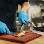 Cozinheiro com avental preto e luvas nitrílicas azuis fatiando frango grelhado sobre tábua vermelha com pegador, ao lado de frigideira com legumes e prato dourado em bancada de madeira