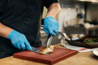Cozinheiro com avental preto e luvas nitrílicas azuis fatiando frango grelhado sobre tábua vermelha com pegador, ao lado de frigideira com legumes e prato dourado em bancada de madeira