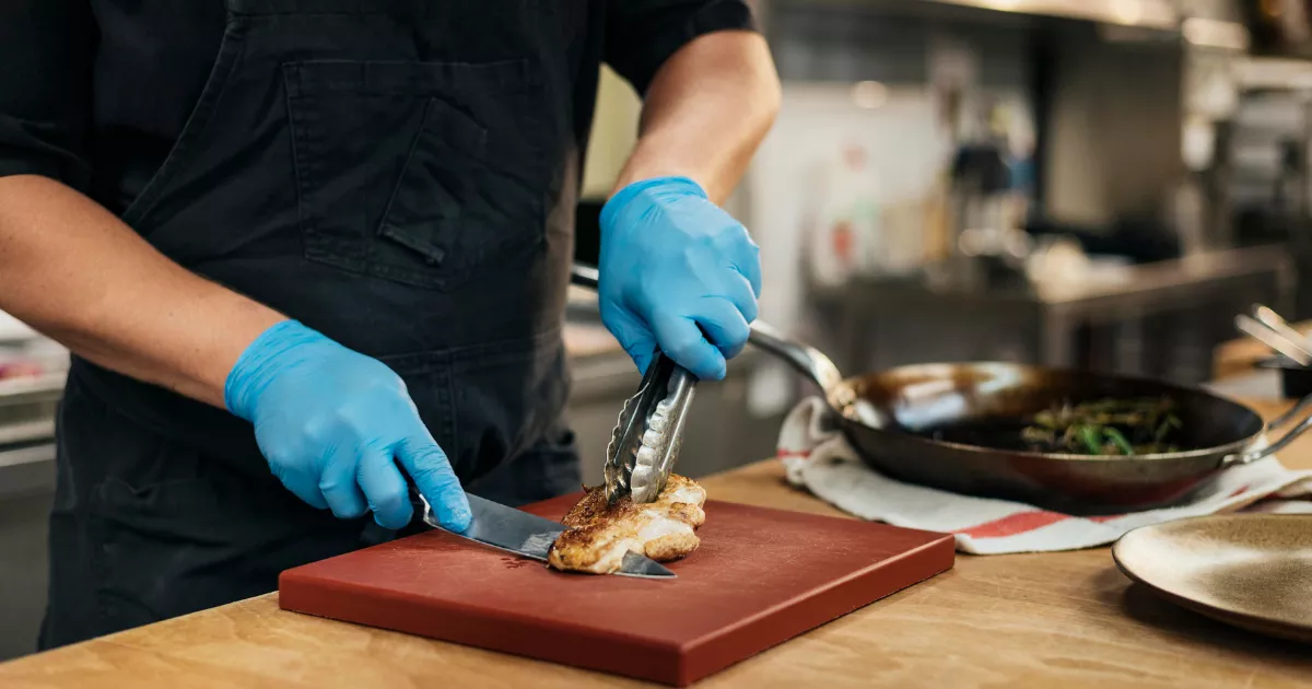 Cozinheiro com avental preto e luvas nitrílicas azuis fatiando frango grelhado sobre tábua vermelha com pegador, ao lado de frigideira com legumes e prato dourado em bancada de madeira
