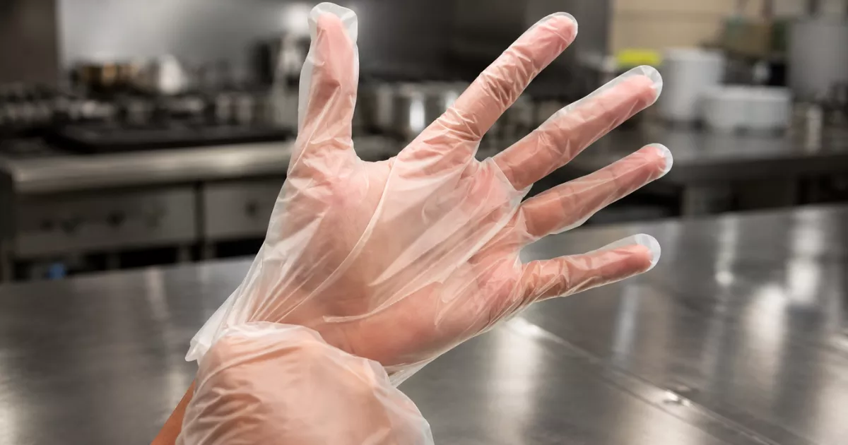 Mão com luva plástica descartável ultrafina e transparente aberta em direção à câmera, com bancada e equipamentos de cozinha profissional desfocados ao fundo