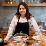 Empreendedora de avental preto apoiada no balcão de madeira de uma cafeteria, com produtos de panificação em redomas de vidro à frente e terminal de caixa ao lado, no contexto de investimento no ramo alimentício
