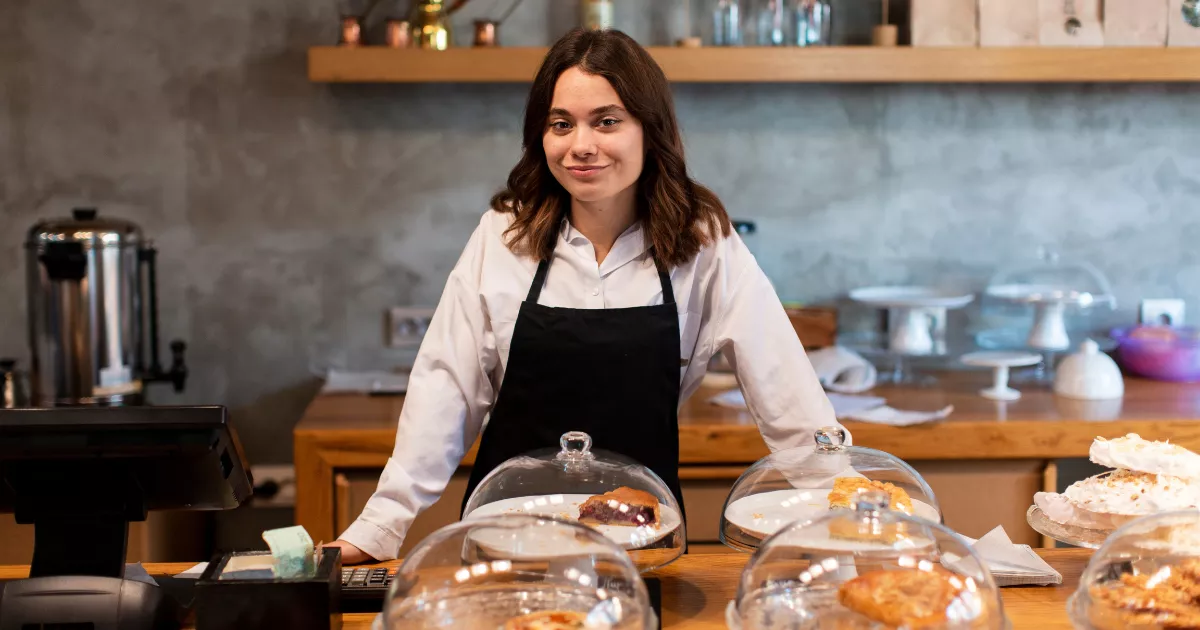 Empreendedora de avental preto apoiada no balcão de madeira de uma cafeteria, com produtos de panificação em redomas de vidro à frente e terminal de caixa ao lado, no contexto de investimento no ramo alimentício