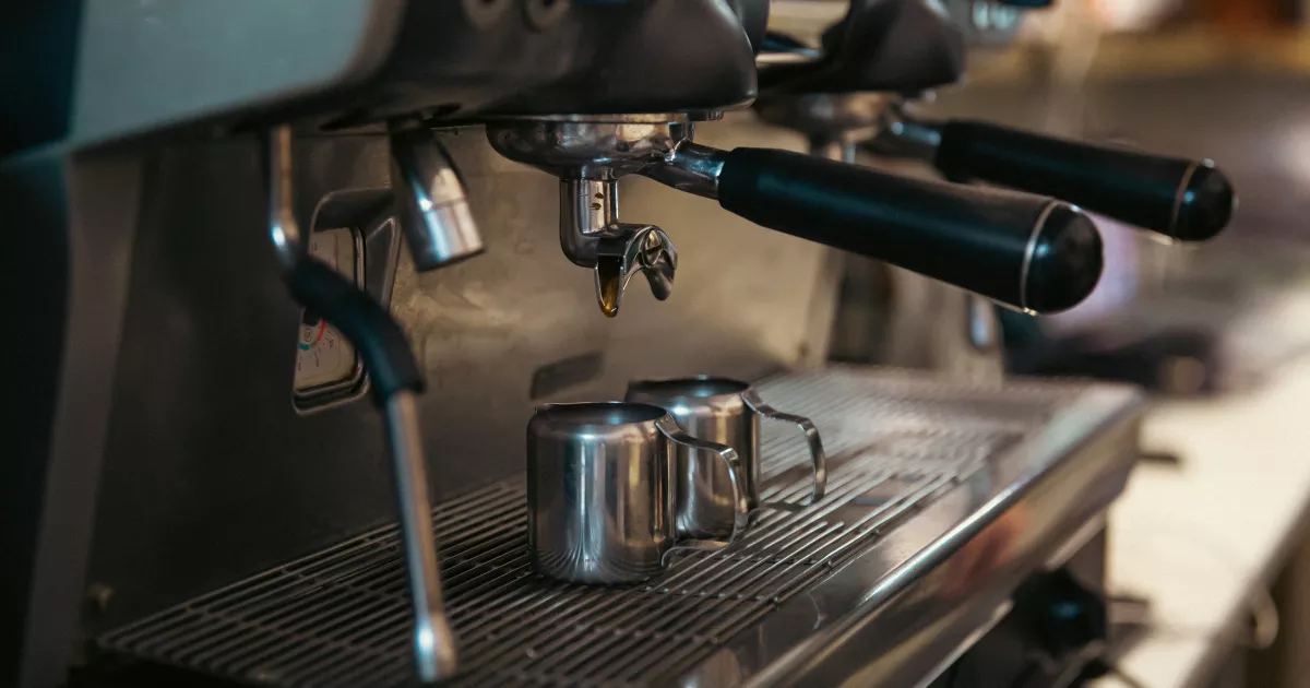 Máquina de espresso profissional de dois grupos extraindo café em pitchers de aço inox sobre a grade metálica do equipamento, em cafeteria com iluminação quente