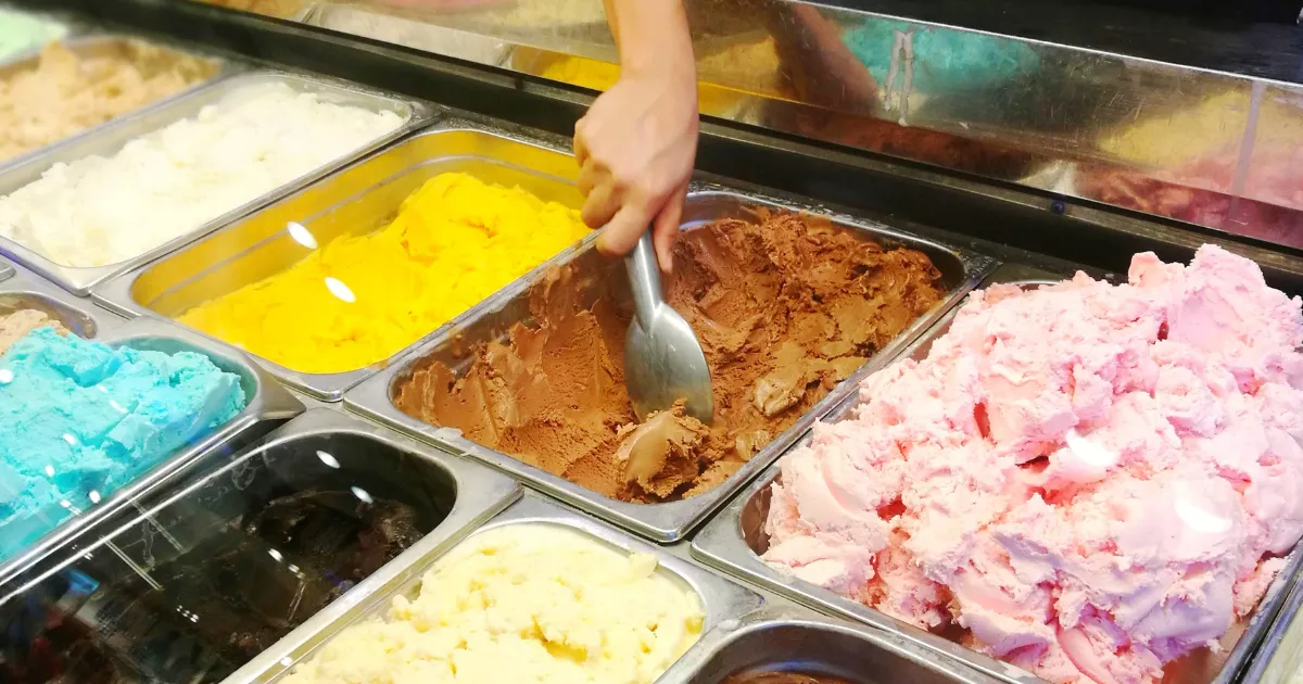 Mão servindo sorvete de chocolate com colher de inox em expositor refrigerado com cubas de sorvete em várias cores, incluindo amarelo, rosa, azul e creme