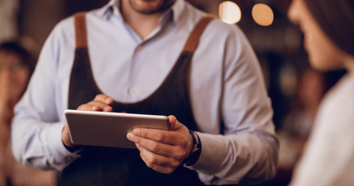 
Garçom de avental marrom e camisa social segura tablet nas duas mãos enquanto atende cliente em restaurante com iluminação quente ao fundo