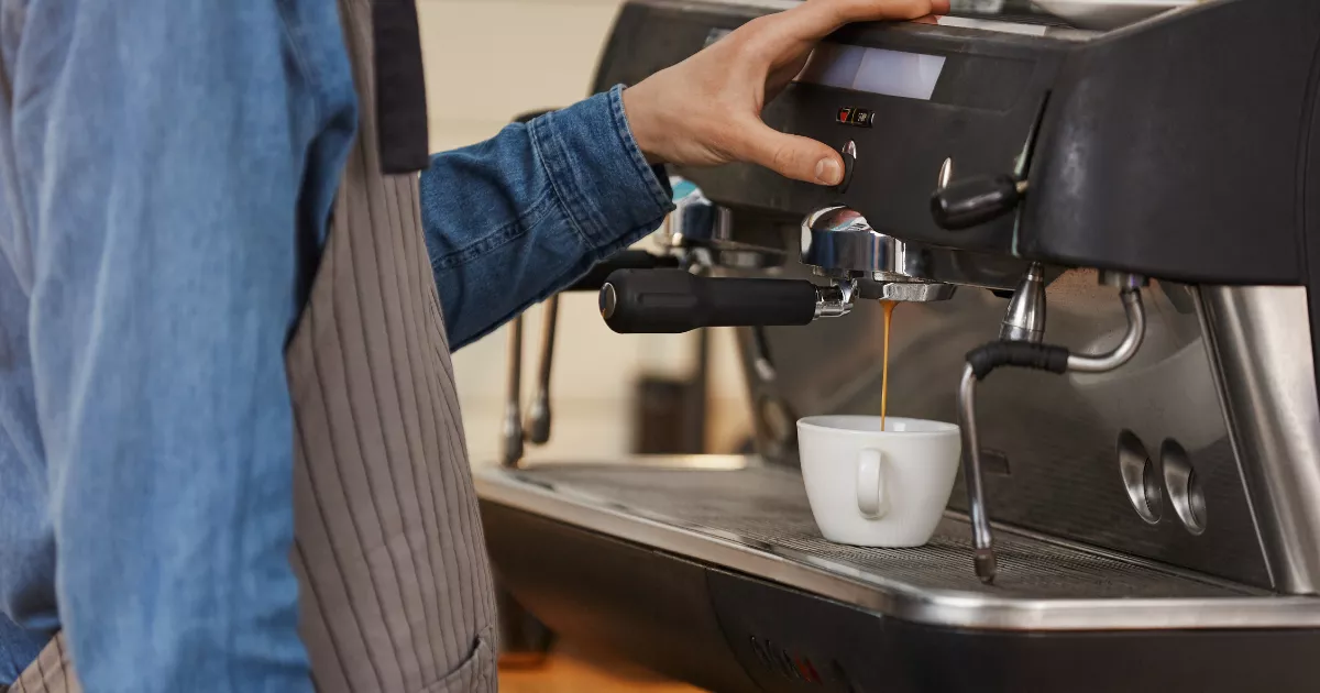 Barista de avental cinza e camisa jeans operando máquina de espresso profissional escura enquanto extrai café em xícara branca, com painel de controles visível