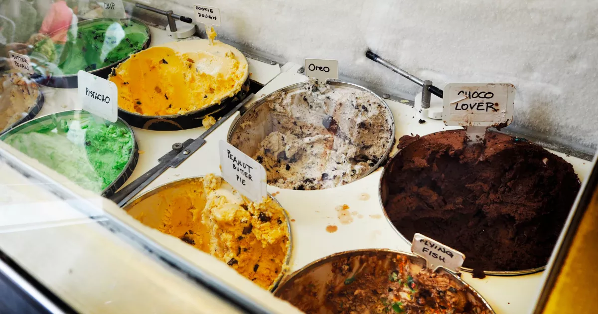 Expositor refrigerado de sorveteria com potes de sorvete em sabores variados como pistache, oreo, cookie dough e chocolate, cada um identificado por plaquinhas escritas à mão