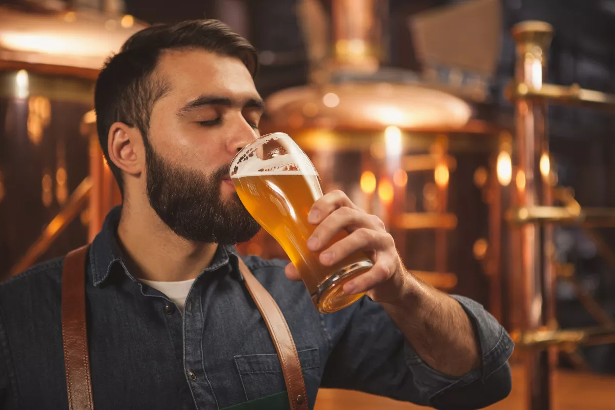 Homem de barba com avental de couro bebe cerveja artesanal clara em taça dentro de uma cervejaria com tanques de cobre ao fundo