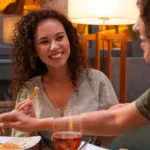 Duas mulheres jovens jantando juntas em restaurante ao ar livre à noite, compartilhando petiscos e taças de vinho rosé, com iluminação quente ao fundo
