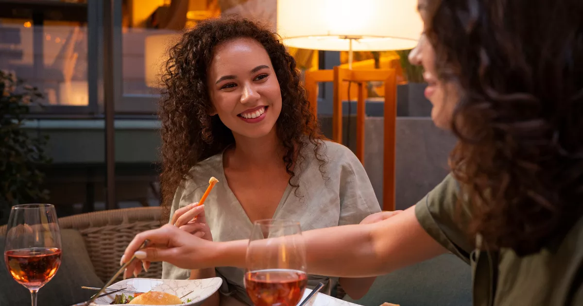 Duas mulheres jovens jantando juntas em restaurante ao ar livre à noite, compartilhando petiscos e taças de vinho rosé, com iluminação quente ao fundo
