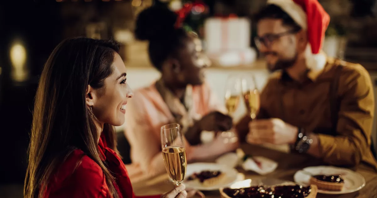 Grupo de três amigos comemorando em jantar de Natal em restaurante, com mulher de blusa vermelha segurando taça de espumante em primeiro plano e homem com gorro de Papai Noel ao fundo