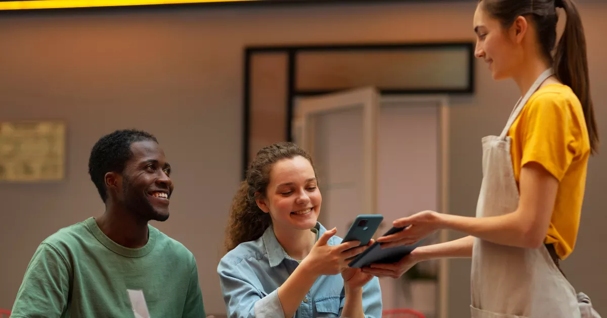 Garçonete de avental bege mostrando tablet para casal de clientes sentados em restaurante, com cliente do sexo feminino usando celular para escanear ou visualizar cardápio digital