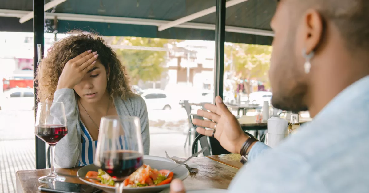 Mulher de cabelo cacheado com a mão na testa, expressão de preocupação, sentada à mesa de restaurante ao ar livre com taças de vinho e prato de salada