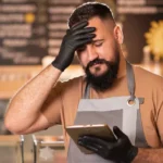 Dono de restaurante de avental e luvas pretas com a mão na testa, olhando o celular com expressão de frustração, atrás do balcão da confeitaria