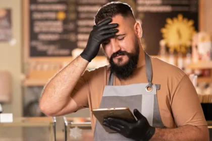 Dono de restaurante de avental e luvas pretas com a mão na testa, olhando o celular com expressão de frustração, atrás do balcão da confeitaria