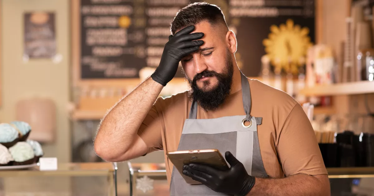 Dono de restaurante de avental e luvas pretas com a mão na testa, olhando o celular com expressão de frustração, atrás do balcão da confeitaria