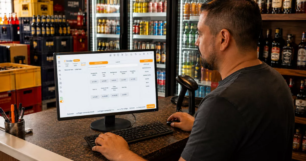 Dono de distribuidora opera o ControleNaMão em monitor de balcão, cercado por geladeiras, bebidas e engradados organizados.