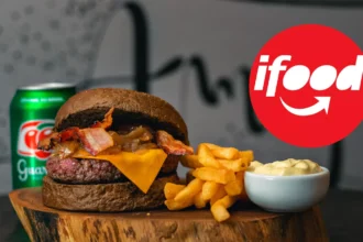 Hambúrguer com bacon, cebola caramelizada e cheddar no pão escuro, batata frita, maionese e lata de guaraná sobre tronco de madeira e logotipo do iFood, um combo típico de delivery