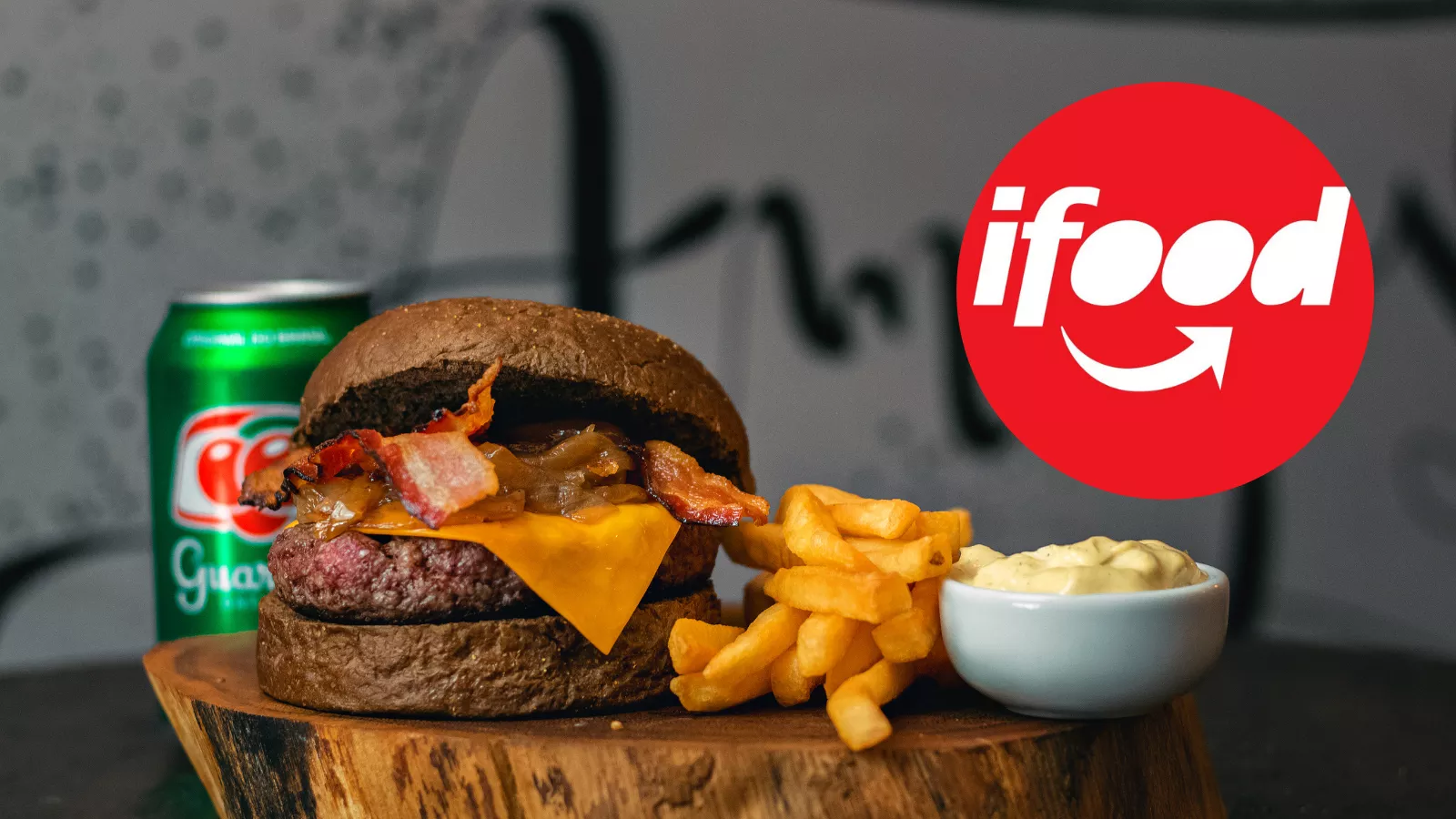 Hambúrguer com bacon, cebola caramelizada e cheddar no pão escuro, batata frita, maionese e lata de guaraná sobre tronco de madeira e logotipo do iFood, um combo típico de delivery
