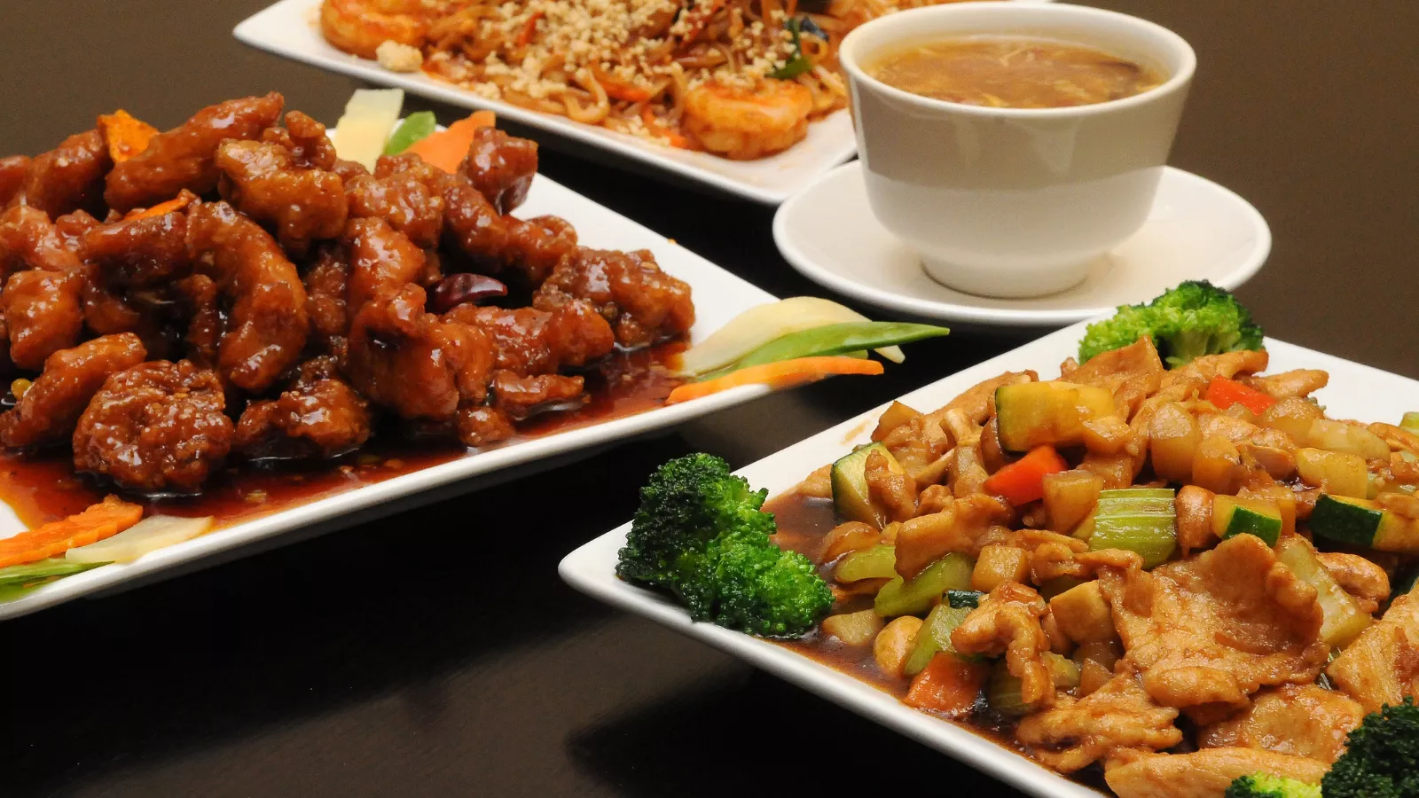 Pratos de comida chinesa sobre mesa escura: pedaços empanados ao molho agridoce, frango salteado com legumes e brócolis, macarrão com camarão e tigela de sopa, opções de combo