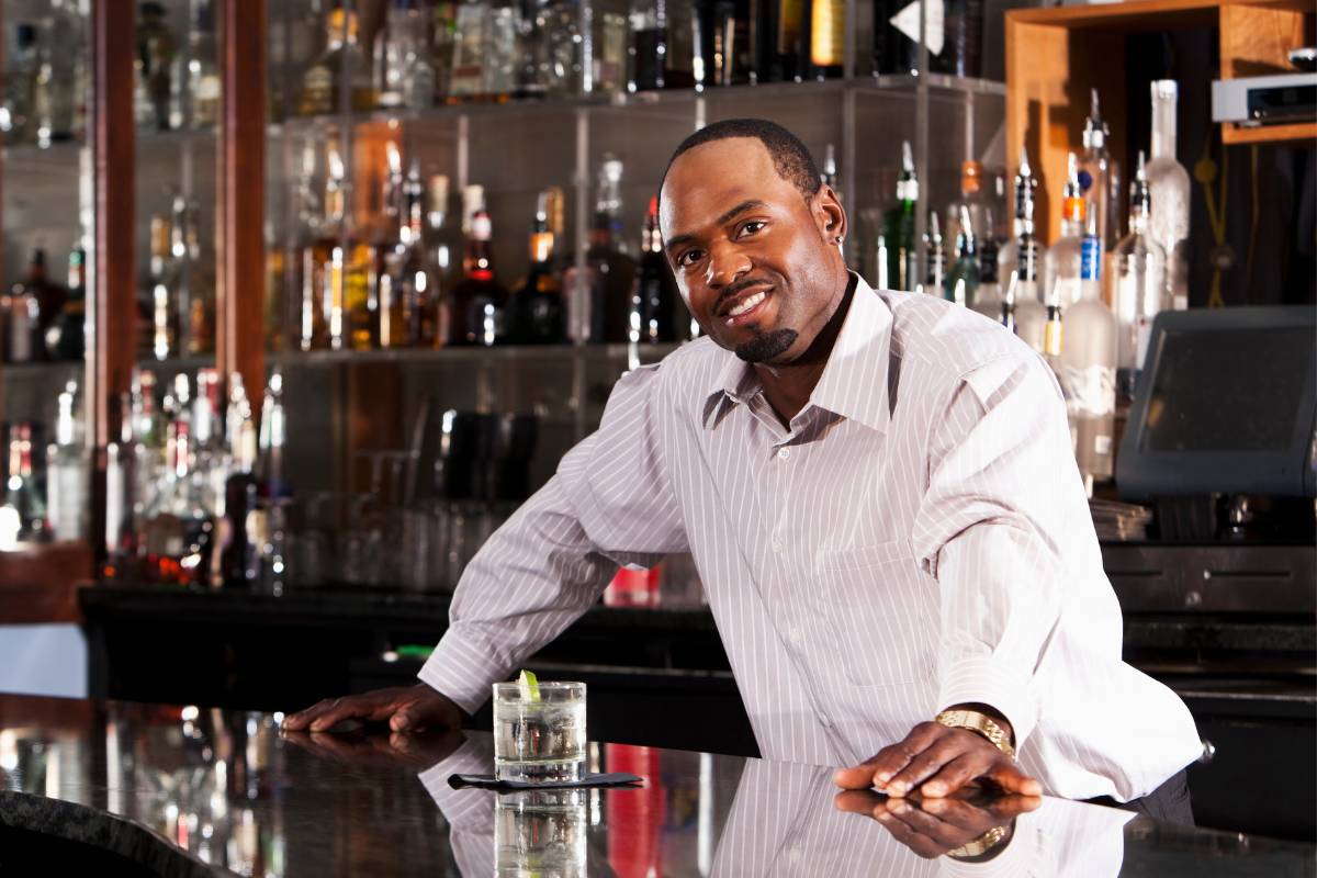 Bartender confiante encostado no balcão de um bar, sorrindo com um drinque na frente.