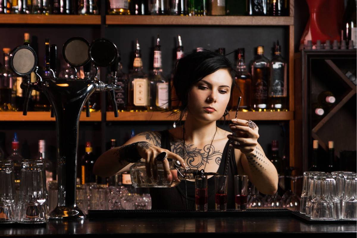 Bartender tatuada preparando drinques atrás do balcão de um bar.